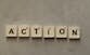 Flat lay of Scrabble tiles spelling 'Action' on a gray background, surrounded by random letters.
